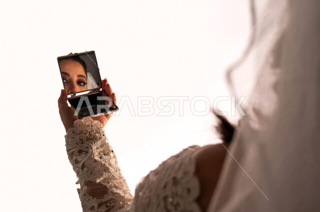 بورتريه بصورة مقربة لإنعكاس صورة عروسة عربية خليجية سعودية جميلة في مرآة صغيرة ، أجمل اطلالة لعروسة عربية سعودية خليجية في يوم زفافها ، ترتدي بدلة الزفاف البيضاء ، ليلة العمر ، مناسبات وأفراح ، خلفية بيضاء