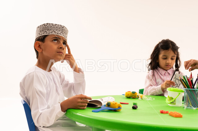 Expressions and gestures of thinking and absent-mindedness, doing fun activities, strengthening artistic skills through shaping, educational sessions and classes, portrait of an Arab Gulf Omani student wearing a keema and a dishdasha playing with clay and colored paste with his friends, developing mental and intellectual abilities, white background