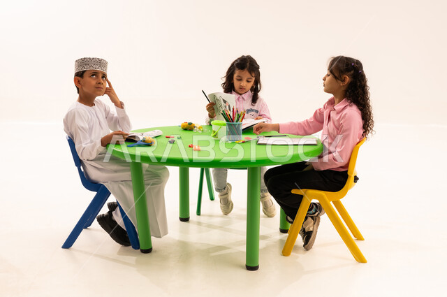Expressions and gestures of thinking and absent-mindedness, doing fun activities, strengthening artistic skills through shaping, educational sessions and classes, portrait of an Arab Gulf Omani student wearing a keema and a dishdasha playing with clay and colored paste with his friends, developing mental and intellectual abilities, white background