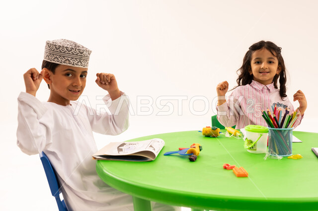 Raising hands with expressions of joy and enthusiasm, doing fun activities, strengthening artistic skills through shaping, educational sessions and classes, portrait of an Arab Gulf Omani student wearing a kummah and a dishdasha playing with clay and colored paste with his friends, developing mental and intellectual abilities, white background