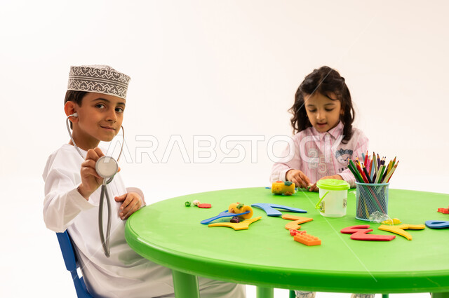 Choosing and determining a future profession from an early age, introducing children in kindergarten to the concept of medicine and health care, a portrait of an Arab Gulf Omani boy wearing a dishdasha and a kuma, holding a stethoscope in his hand, playing with the profession of medicine alongside his friends, white background