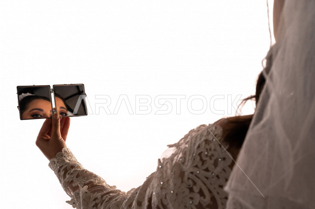 بورتريه بصورة مقربة لإنعكاس صورة عروسة عربية خليجية سعودية جميلة في مرآة صغيرة ، أجمل اطلالة لعروسة عربية سعودية خليجية في يوم زفافها ، ترتدي بدلة الزفاف البيضاء ، ليلة العمر ، مناسبات وأفراح ، خلفية بيضاء