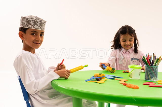 Doing fun and entertaining skills and activities, developing artistic skills through shaping, educational sessions and classes, a portrait of an Arab Gulf Omani student wearing a kummah and a dishdasha playing with clay and colored paste with his friends, developing mental and intellectual abilities, white background