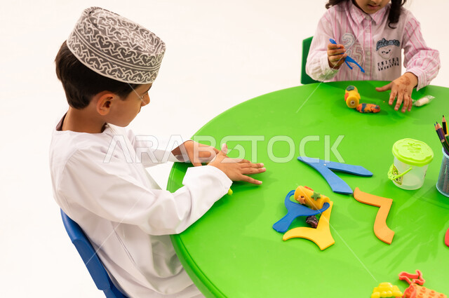 Doing fun and entertaining skills and activities, developing artistic skills through shaping, educational sessions and classes, a portrait of an Arab Gulf Omani student wearing a kummah and a dishdasha playing with clay and colored paste with his friends, developing mental and intellectual abilities, white background