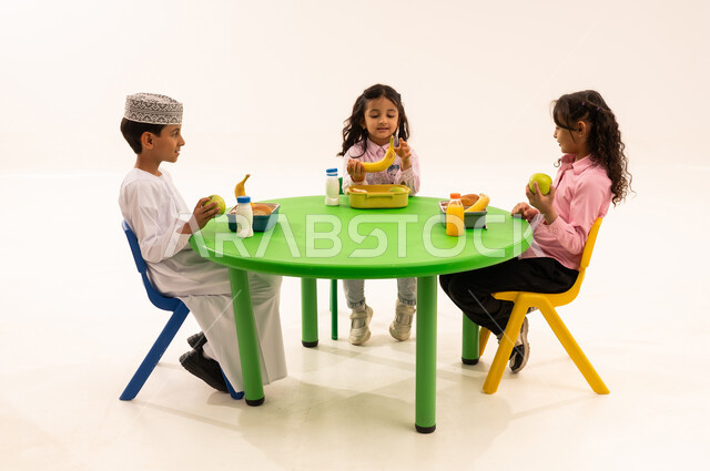 أسس الغذاء الصحي السليم، تناول الطعام في وقت الفراغ، فترة الاستراحة في الروضة، بورتريه لولد عربي خليجي عماني يرتدي الدشداشة والكمة يقوم بتناول الطعام الصحي وقت الفسحة المدرسية مع اصدقاءه، المدارس والروضات العمانية، خلفية بيضاء