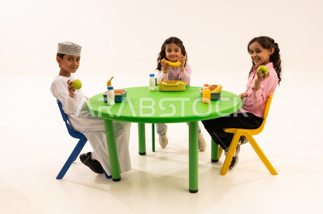 Eating during free time, the foundations of healthy eating, recess in kindergarten, a portrait of an Arab Gulf Omani boy wearing a dishdasha and a kummah eating healthy food during school break with his friends, Omani schools and kindergartens, white background