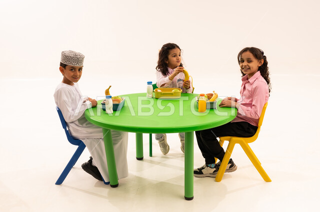 Foundations of healthy eating, eating during free time, recess in kindergarten, portrait of an Arab Gulf Omani boy wearing a dishdasha and a kummah eating healthy food during school recess with his friends, Omani schools and kindergartens, white background