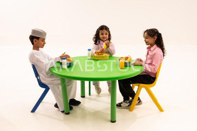 تناول الطعام في وقت الفراغ، أسس الغذاء الصحي السليم، فترة الاستراحة في الروضة، بورتريه لولد عربي خليجي عماني يرتدي الدشداشة والكمة يقوم بتناول الطعام الصحي وقت الفسحة المدرسية مع اصدقاءه، المدارس والروضات العمانية، خلفية بيضاء