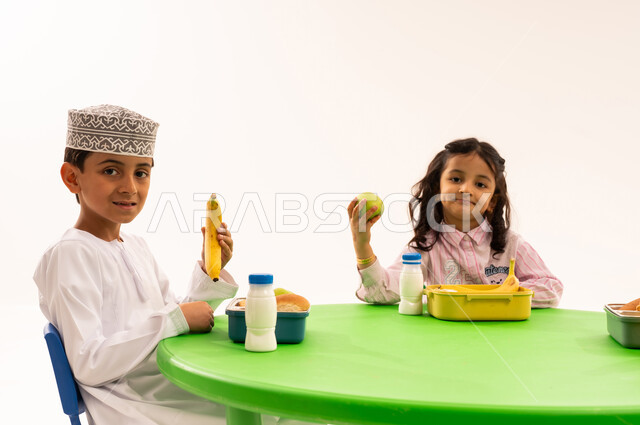 أسس الغذاء الصحي السليم، تناول الطعام في وقت الفراغ، فترة الاستراحة في الروضة، بورتريه لولد عربي خليجي عماني يرتدي الدشداشة والكمة يقوم بتناول الطعام الصحي وقت الفسحة المدرسية مع اصدقاءه، المدارس والروضات العمانية، خلفية بيضاء