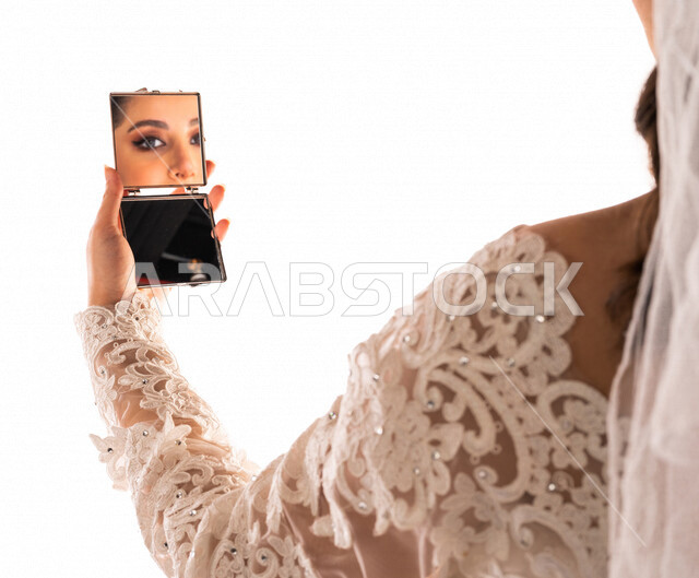 بورتريه بصورة مقربة لإنعكاس صورة عروسة عربية خليجية سعودية جميلة في مرآة صغيرة ، أجمل اطلالة لعروسة عربية سعودية خليجية في يوم زفافها ، ترتدي بدلة الزفاف البيضاء ، ليلة العمر ، مناسبات وأفراح ، خلفية بيضاء