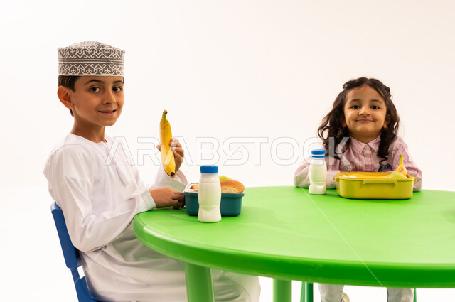 أسس الغذاء الصحي السليم، تناول الطعام في وقت الفراغ، فترة الاستراحة في الروضة، بورتريه لولد عربي خليجي عماني يرتدي الدشداشة والكمة يقوم بتناول الطعام الصحي وقت الفسحة المدرسية مع اصدقاءه، المدارس والروضات العمانية، خلفية بيضاء