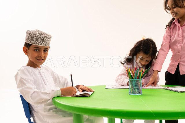 تعزيز المهارات الفنية بالرسم والتلوين، الجلسات والحصص التعليمية في الروضات العمانية، بورتريه لفتى عربي خليجي عماني يرتدي الكمة والدشداشة يقوم بتلوين الرسومات بايماءات الاندماج مع اصدقاءه، القيام بالمهارات و الأنشطة المختلفة لتنمية القدرات العقلية والذهنية، خلفية بيضاء