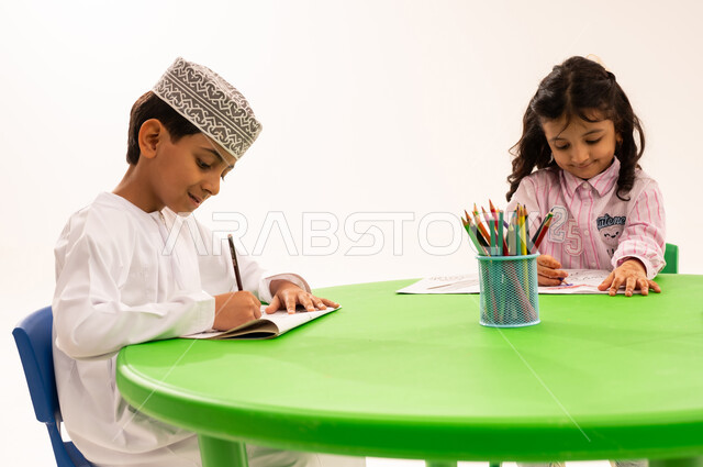 تعزيز المهارات الفنية بالرسم والتلوين، الجلسات والحصص التعليمية في الروضات العمانية، بورتريه لفتى عربي خليجي عماني يرتدي الكمة والدشداشة يقوم بتلوين الرسومات بايماءات الاندماج مع اصدقاءه، القيام بالمهارات و الأنشطة المختلفة لتنمية القدرات العقلية والذهنية، خلفية بيضاء