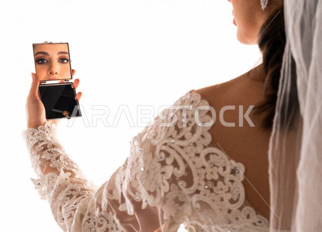 بورتريه بصورة مقربة لإنعكاس صورة عروسة عربية خليجية سعودية جميلة في مرآة صغيرة ، أجمل اطلالة لعروسة عربية سعودية خليجية في يوم زفافها ، ترتدي بدلة الزفاف البيضاء ، ليلة العمر ، مناسبات وأفراح ، خلفية بيضاء