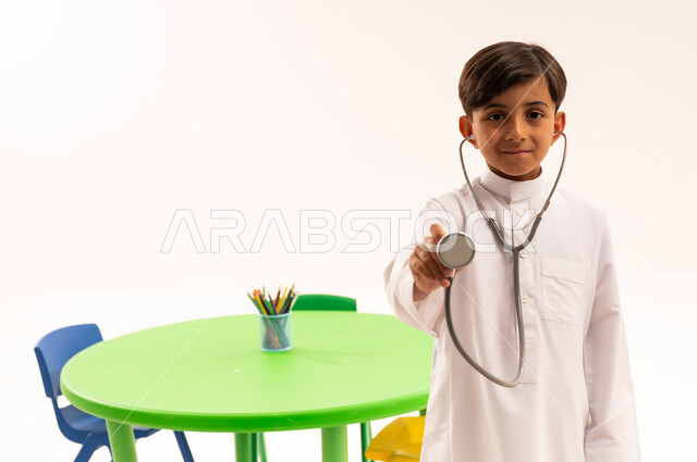 Choosing and determining a future profession from an early age, introducing children in kindergarten to the concept of medicine and healthcare, a portrait of a Saudi Arabian Gulf boy wearing traditional clothing holding a stethoscope while playing with the profession of medicine, white background