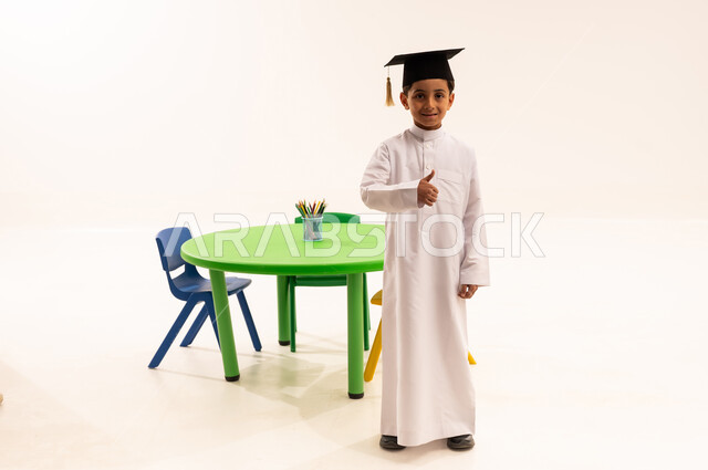 النجاح والتفوق في المرحلة الدراسية، بورتريه لولد عربي خليجي سعودي يرتدي الثوب التقليدي وقبعة التخرج من ينظز الى الكاميرا بتعابير السرور والسعادة، انهاء المرحلة التعليمية الاولى، الجسم بالطول الكامل، خلفية بيضاء