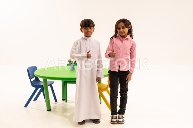 الاستعداد لبدء عام دراسي جديد، المدارس والروضات في السعودية، ايماءات وتعابير السرور والسعادة لبداية الفصل الدراسي، بورتريه لولد عربي خليجي سعودي يرتدي الثوب التقليدي يقف باستقامة ينظر الى الكاميرا بجانب صديقته، الطول الكامل للجسم، خلفية بيضاء