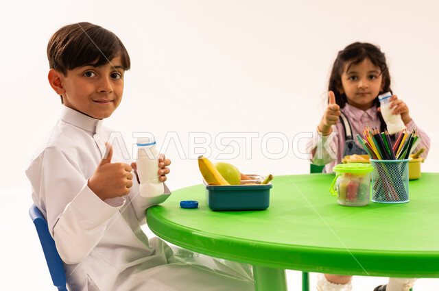 أسس الغذاء الصحي السليم، وقت الفراغ وتناول الطعام، فترة الاستراحة في الروضة، بورتريه لاولاد عرب خليجيين سعوديين يرتدون الزي الكاجوال يتناولون الطعام الصحي وقت الفسحة المدرسية في إحدى رياض الأطفال في السعودية، خلفية بيضاء