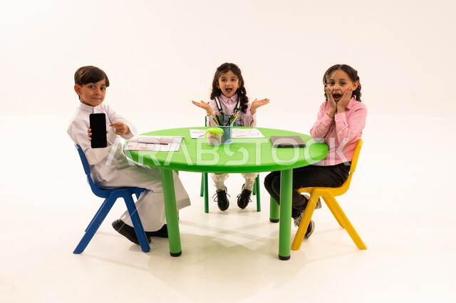 Empty black screen display, entertaining activity, enhancing students' artistic creativity, portrait of a Saudi Arabian Gulf student wearing traditional clothing holding a mobile phone sitting next to his friends, drawing, coloring, and using modern technical devices, white background