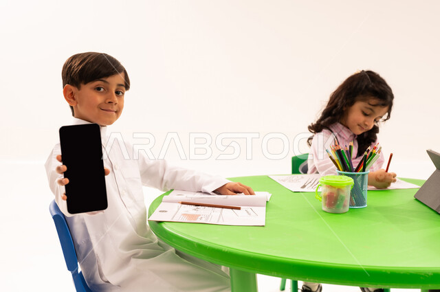 Empty black screen display, entertaining activity, enhancing students' artistic creativity, portrait of a Saudi Arabian Gulf student wearing traditional clothing holding a mobile phone sitting next to his friends, drawing, coloring, and using modern technical devices, white background