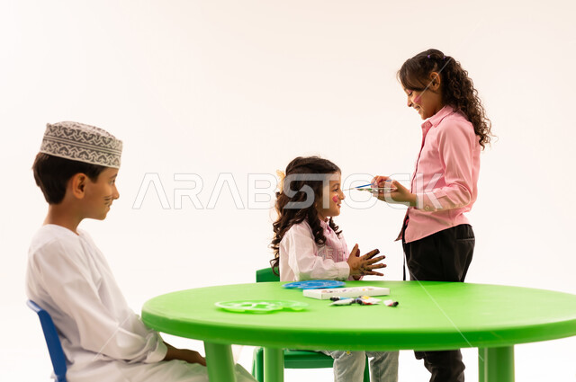 تعابير وايماءات السرور والفرح، الرسم على وجوه الاولاد بالالوان الصحية، قضاء أوقات ممتعة ومفيدة في الروضة، بورتريه لطلاب عرب خليجيين عمانين يقومون بأنشطة ترفيهيه مسلية داخل الفصل الدراسي، خلفية بيضاء