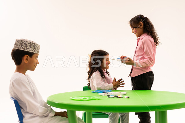 تعابير وايماءات السرور والفرح، الرسم على وجوه الاولاد بالالوان الصحية، قضاء أوقات ممتعة ومفيدة في الروضة، بورتريه لطلاب عرب خليجيين عمانين يقومون بأنشطة ترفيهيه مسلية داخل الفصل الدراسي، خلفية بيضاء