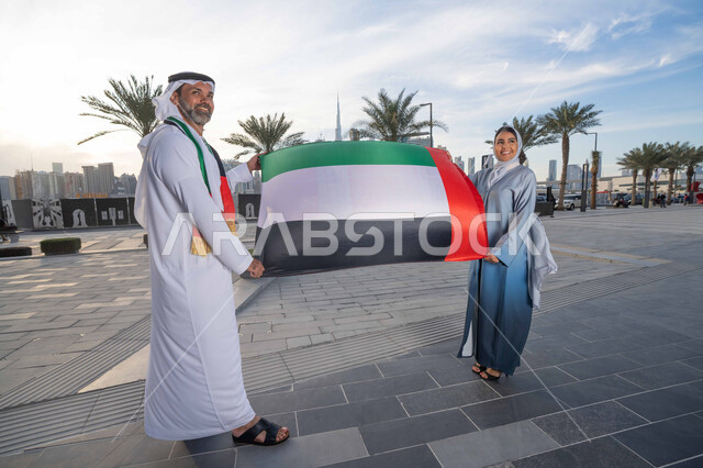 ذكرى استقلال دولة الإمارات العربية المتحدة، اليوم الوطني الإماراتي 2 ديسمبر، رجل عربي خليجي اماراتي يرتدي الكندورة والغترة يحتفل بالعيد الوطني برفقة زوجته في النهار، التجول في الشوارع بايماءات السعادة والفخر والاعتزاز، حب الوطن والانتماء اليه، رفع علم وراية الوطن