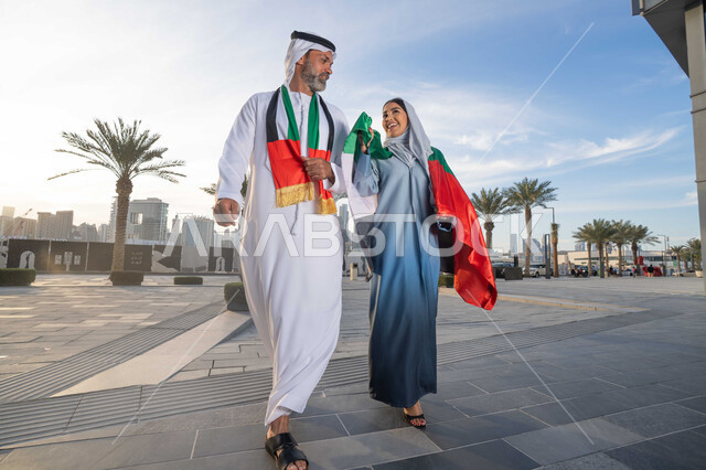 المشي في الشوارع بايماءات السعادة والفخر والاعتزاز، حب الوطن والانتماء اليه، علم وراية الوطن، ذكرى استقلال دولة الإمارات العربية المتحدة، اليوم الوطني الإماراتي 2 ديسمبر، رجل عربي خليجي اماراتي يرتدي الكندورة والغترة يحتفل بالعيد الوطني برفقة زوجته في النهار