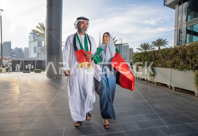 علم وراية الوطن، ذكرى استقلال دولة الإمارات العربية المتحدة، اليوم الوطني الإماراتي 2 ديسمبر، رجل عربي خليجي اماراتي يرتدي الكندورة والغترة يحتفل بالعيد الوطني برفقة زوجته في النهار، التجول في الشوارع بايماءات السعادة والفخر والاعتزاز، حب الوطن والانتماء اليه