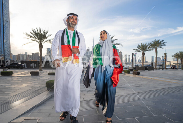 التجول في الشوارع بايماءات السعادة والفخر والاعتزاز، حب الوطن والانتماء اليه، علم وراية الوطن، ذكرى استقلال دولة الإمارات العربية المتحدة، اليوم الوطني الإماراتي 2 ديسمبر، رجل عربي خليجي اماراتي يرتدي الكندورة والغترة يحتفل بالعيد الوطني برفقة زوجته في النهار