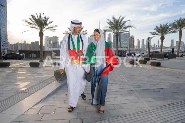 المشي في الشوارع بايماءات السعادة والفخر والاعتزاز، حب الوطن والانتماء اليه، علم وراية الوطن، ذكرى استقلال دولة الإمارات العربية المتحدة، اليوم الوطني الإماراتي 2 ديسمبر، رجل عربي خليجي اماراتي يرتدي الكندورة والغترة يحتفل بالعيد الوطني برفقة زوجته في النهار