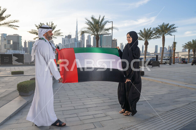 علم وراية الوطن، ذكرى استقلال دولة الإمارات العربية المتحدة، اليوم الوطني الإماراتي 2 ديسمبر، رجل عربي خليجي اماراتي يرتدي الكندورة والغترة يحتفل بالعيد الوطني برفقة زوجته في النهار، التجول في الشوارع بايماءات السعادة والفخر والاعتزاز، حب الوطن والانتماء اليه