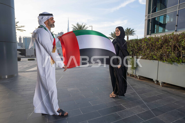 علم وراية الوطن، ذكرى استقلال دولة الإمارات العربية المتحدة، اليوم الوطني الإماراتي 2 ديسمبر، رجل عربي خليجي اماراتي يرتدي الكندورة والغترة يحتفل بالعيد الوطني برفقة زوجته في النهار، التجول في الشوارع بايماءات السعادة والفخر والاعتزاز، حب الوطن والانتماء اليه