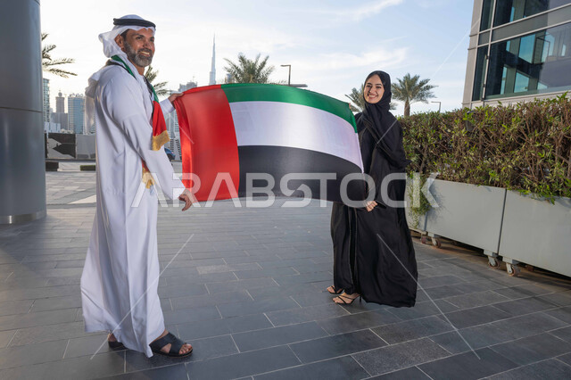 ذكرى استقلال دولة الإمارات العربية المتحدة، اليوم الوطني الإماراتي 2 ديسمبر، رجل عربي خليجي اماراتي يرتدي الكندورة والغترة يحتفل بالعيد الوطني برفقة زوجته في النهار، التجول في الشوارع بايماءات السعادة والفخر والاعتزاز، حب الوطن والانتماء اليه، رفع علم وراية الوطن