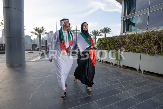 التجول في الشوارع بايماءات السعادة والفخر والاعتزاز، حب الوطن والانتماء اليه، علم وراية الوطن، ذكرى استقلال دولة الإمارات العربية المتحدة، اليوم الوطني الإماراتي 2 ديسمبر، رجل عربي خليجي اماراتي يرتدي الكندورة والغترة يحتفل بالعيد الوطني برفقة زوجته في النهار