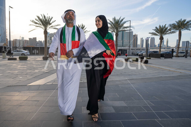 المشي في الشوارع بايماءات السعادة والفخر والاعتزاز، حب الوطن والانتماء اليه، علم وراية الوطن، ذكرى استقلال دولة الإمارات العربية المتحدة، اليوم الوطني الإماراتي 2 ديسمبر، رجل عربي خليجي اماراتي يرتدي الكندورة والغترة يحتفل بالعيد الوطني برفقة زوجته في النهار