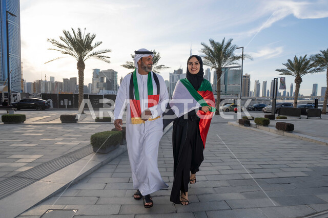 التجول في الشوارع بايماءات السعادة والفخر والاعتزاز، حب الوطن والانتماء اليه، علم وراية الوطن، ذكرى استقلال دولة الإمارات العربية المتحدة، اليوم الوطني الإماراتي 2 ديسمبر، رجل عربي خليجي اماراتي يرتدي الكندورة والغترة يحتفل بالعيد الوطني برفقة زوجته في النهار