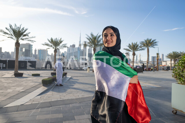 اليوم الوطني الإماراتي 2 ديسمبر،  الاحتفال بذكرى استقلال دولة الإمارات العربية المتحدة، امراة عربية خليجية اماراتية ترتدي العباءة والحجاب تضع علم الإمارات على كتفيها في النهار، التجول في الشوارع بايماءات السعادة والفخر والاعتزاز، مفهوم حب الوطن والانتماء اليه
