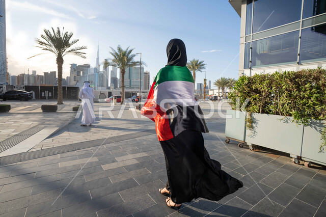 اليوم الوطني الإماراتي 2 ديسمبر،  الاحتفال بذكرى استقلال دولة الإمارات العربية المتحدة، امراة عربية خليجية اماراتية ترتدي العباءة والحجاب تضع علم الإمارات على كتفيها في النهار، التجول في الشوارع بايماءات السعادة والفخر والاعتزاز، مفهوم حب الوطن والانتماء اليه