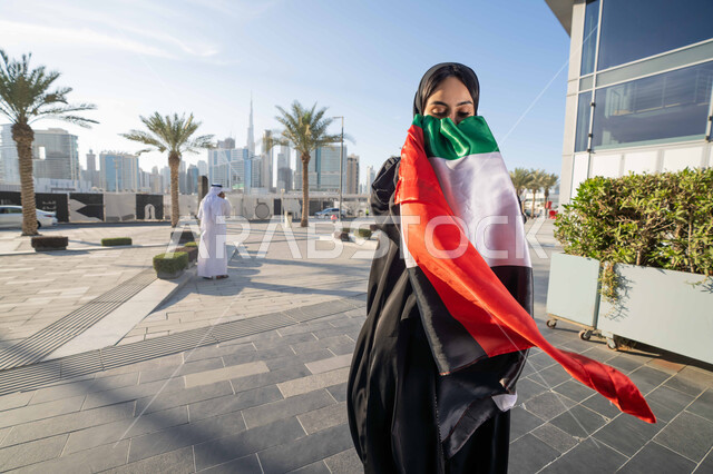 احياء اليوم الوطني الإماراتي 2 ديسمبر،  الاحتفال بذكرى استقلال دولة الإمارات العربية المتحدة، امراة عربية خليجية اماراتية ترتدي العباءة والحجاب تقبل علم الإمارات في النهار، التجول في الشوارع بايماءات السعادة والفخر والاعتزاز، مفهوم حب الوطن والانتماء اليه