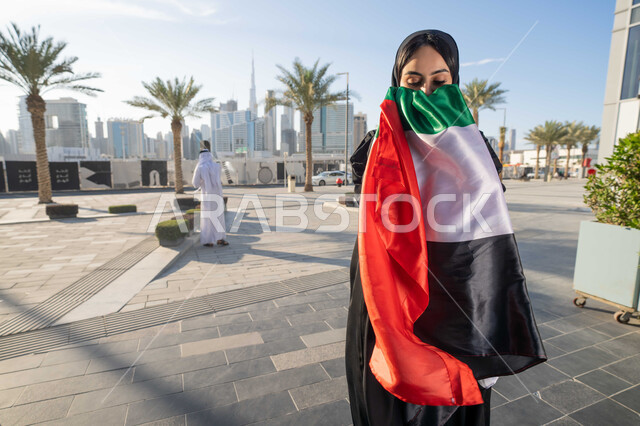 الاحتفال بذكرى استقلال دولة الإمارات العربية المتحدة، امراة عربية خليجية اماراتية ترتدي العباءة والحجاب تقبل علم الإمارات في النهار، التجول في الشوارع بايماءات السعادة والفخر والاعتزاز، مفهوم حب الوطن والانتماء اليه، اليوم الوطني الإماراتي 2 ديسمبر
