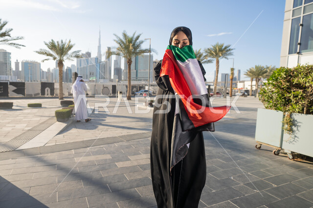 Celebrating the UAE National Day on December 2, celebrating the anniversary of the independence of the United Arab Emirates, an Emirati Gulf Arab woman wearing an abaya and hijab kissing the UAE flag during the day, walking in the streets with gestures of happiness, pride and honor, the concept of love of the homeland and belonging to it