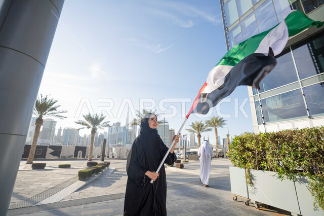 الاحتفال باليوم الوطني الإماراتي 2 ديسمبر، إحياء ذكرى استقلال دولة الإمارات العربية المتحدة، امراة عربية خليجية اماراتية ترتدي العباءة والحجاب ترفع علم الإمارات في النهار، التجول في الشوارع بايماءات السعادة والفخر والاعتزاز، مفهوم حب الوطن والانتماء اليه