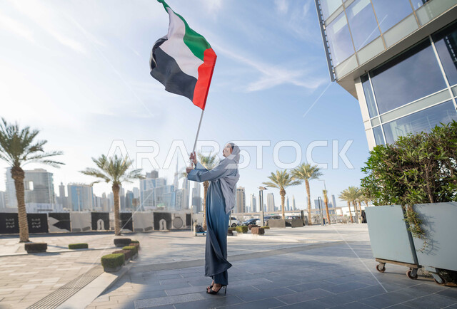 الاحتفال باليوم الوطني الإماراتي 2 ديسمبر، إحياء ذكرى استقلال دولة الإمارات العربية المتحدة، امراة عربية خليجية اماراتية ترتدي العباءة والحجاب ترفع علم الإمارات في النهار، التجول في الشوارع بايماءات السعادة والفخر والاعتزاز، مفهوم حب الوطن والانتماء اليه
