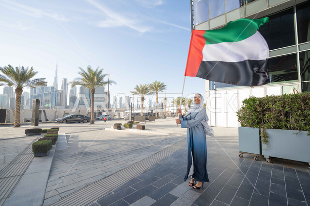 الاحتفال باليوم الوطني الإماراتي 2 ديسمبر، إحياء ذكرى استقلال دولة الإمارات العربية المتحدة، امراة عربية خليجية اماراتية ترتدي العباءة والحجاب ترفع علم الإمارات في النهار، التجول في الشوارع بايماءات السعادة والفخر والاعتزاز، مفهوم حب الوطن والانتماء اليه
