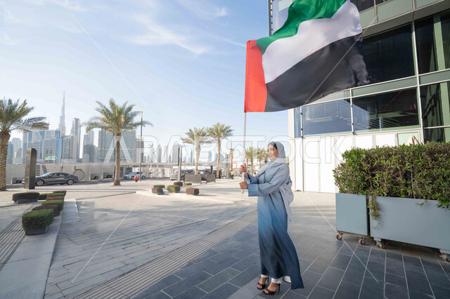 الاحتفال باليوم الوطني الإماراتي 2 ديسمبر، إحياء ذكرى استقلال دولة الإمارات العربية المتحدة، امراة عربية خليجية اماراتية ترتدي العباءة والحجاب ترفع علم الإمارات في النهار، التجول في الشوارع بايماءات السعادة والفخر والاعتزاز، مفهوم حب الوطن والانتماء اليه