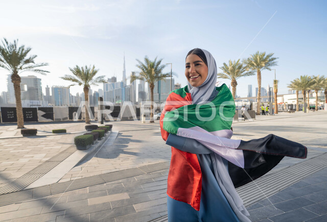 اليوم الوطني الإماراتي 2 ديسمبر،  الاحتفال بذكرى استقلال دولة الإمارات العربية المتحدة، امراة عربية خليجية اماراتية ترتدي العباءة والحجاب تضع علم الإمارات على كتفيها في النهار، التجول في الشوارع بايماءات السعادة والفخر والاعتزاز، مفهوم حب الوطن والانتماء اليه