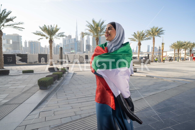 إحياء اليوم الوطني الإماراتي 2 ديسمبر، الاحتفال بذكرى استقلال دولة الإمارات العربية المتحدة، امراة عربية خليجية اماراتية ترتدي العباءة والحجاب تضع علم الإمارات على كتفيها في النهار، التجول في الشوارع بايماءات السعادة والفخر والاعتزاز، مفهوم حب الوطن والانتماء اليه