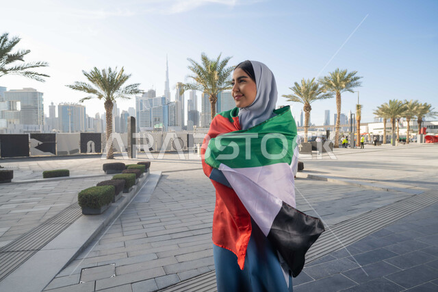 اليوم الوطني الإماراتي 2 ديسمبر،  الاحتفال بذكرى استقلال دولة الإمارات العربية المتحدة، امراة عربية خليجية اماراتية ترتدي العباءة والحجاب تضع علم الإمارات على كتفيها في النهار، التجول في الشوارع بايماءات السعادة والفخر والاعتزاز، مفهوم حب الوطن والانتماء اليه