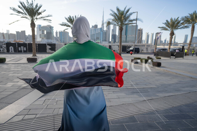 Celebrating the UAE National Day on December 2, celebrating the anniversary of the independence of the United Arab Emirates, an Emirati Gulf Arab woman wearing an abaya and hijab, placing the UAE flag on her shoulders during the day, walking in the streets with gestures of happiness, pride and honor, the concept of love of the homeland and belonging to it
